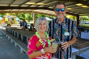 Two attendees enjoying drinks and leis at an outdoor Hawaiian luau celebration under a covered pavilion.