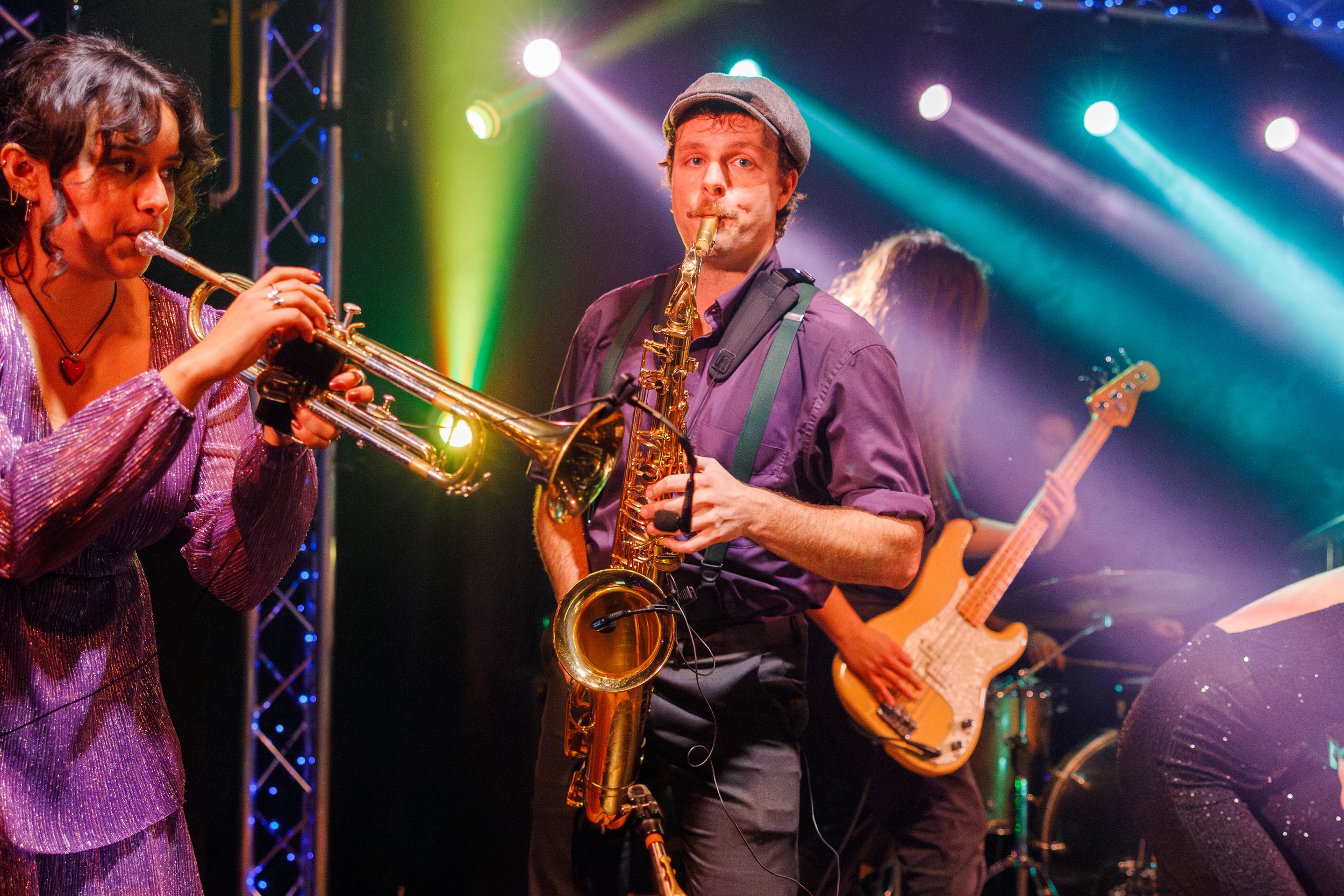 Musicians perform on stage with colorful blue, green and purple lighting creating a dynamic concert atmosphere.