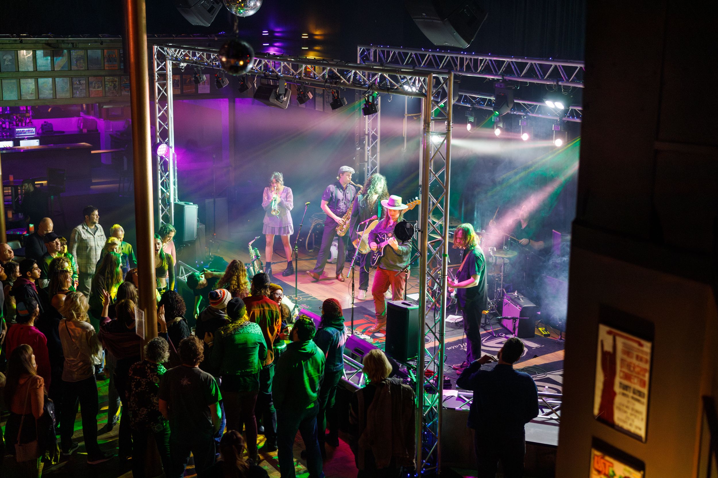 A group of spectators watch a live music performance on stage in a dark venue with colorful purple and green lighting.