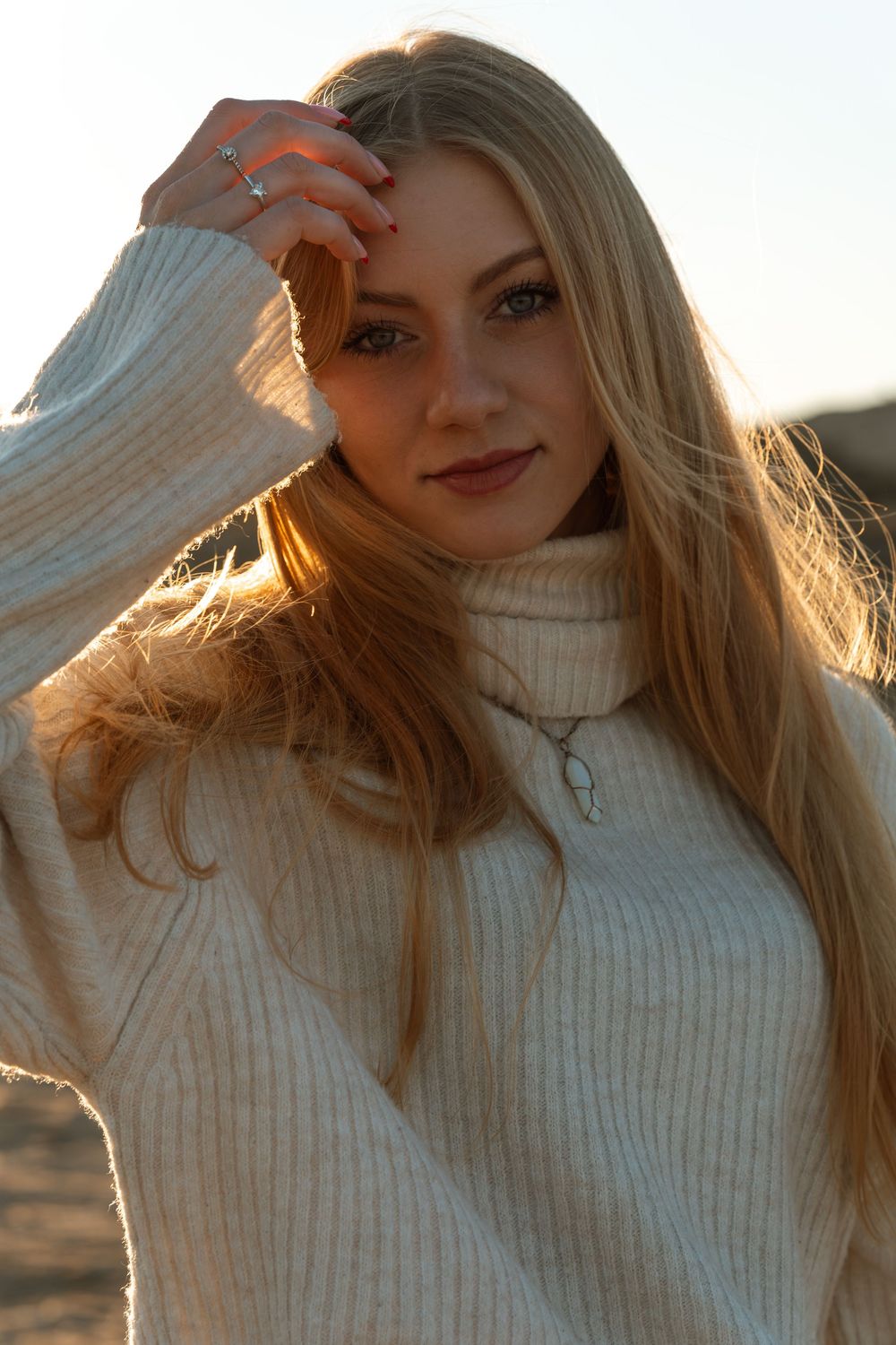 Portret dziewczyny z długimi blond włosami w jasnym swetrze, oświetlonej ciepłym światłem zachodzącego słońca na plaży.
