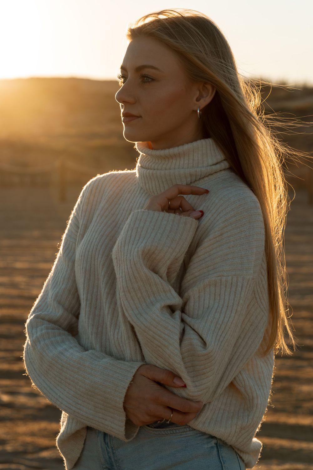 Dziewczyna z długimi blond włosami w jasnym swetrze na plaży o zachodzie słońca.