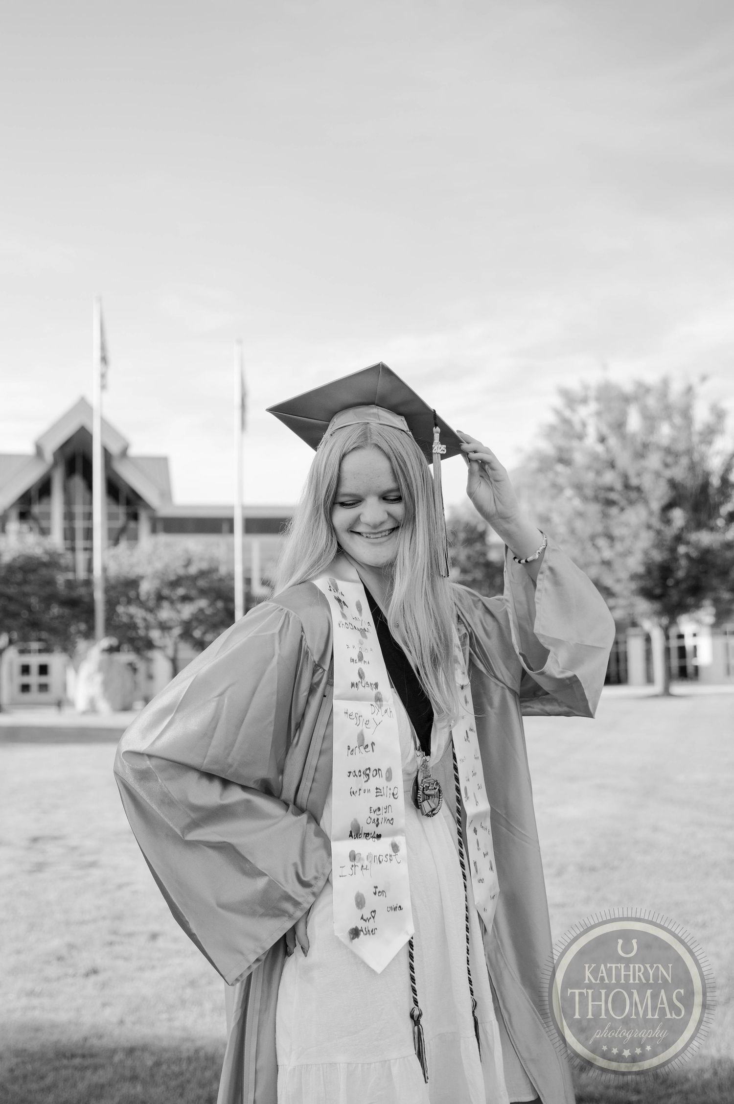 Graduate in bright orange cap and gown holding diploma stands on grassy campus lawn during commencement ceremony.