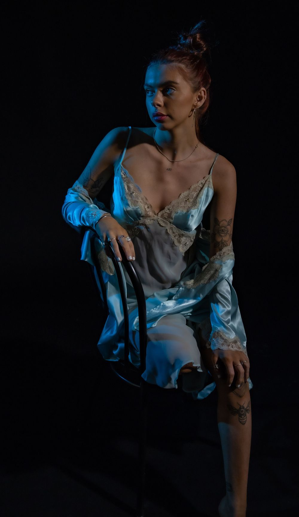 A moody fashion portrait in blue lighting features a model in a delicate white slip dress and sheer robe against dark backdrop.