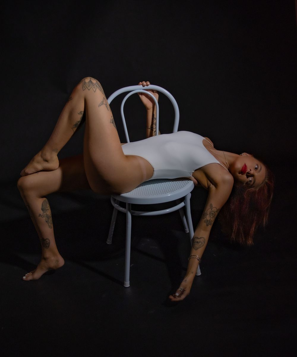 Artistic nude figure poses dramatically on white chair against dark background in studio lighting.