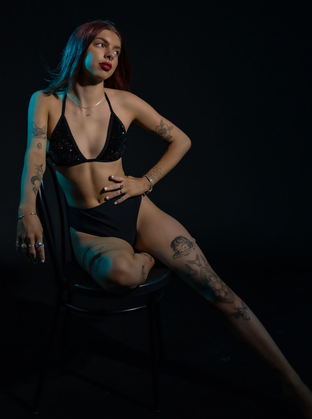 Artistic boudoir photo of a model in black lingerie posing on a chair against dark background with blue lighting.