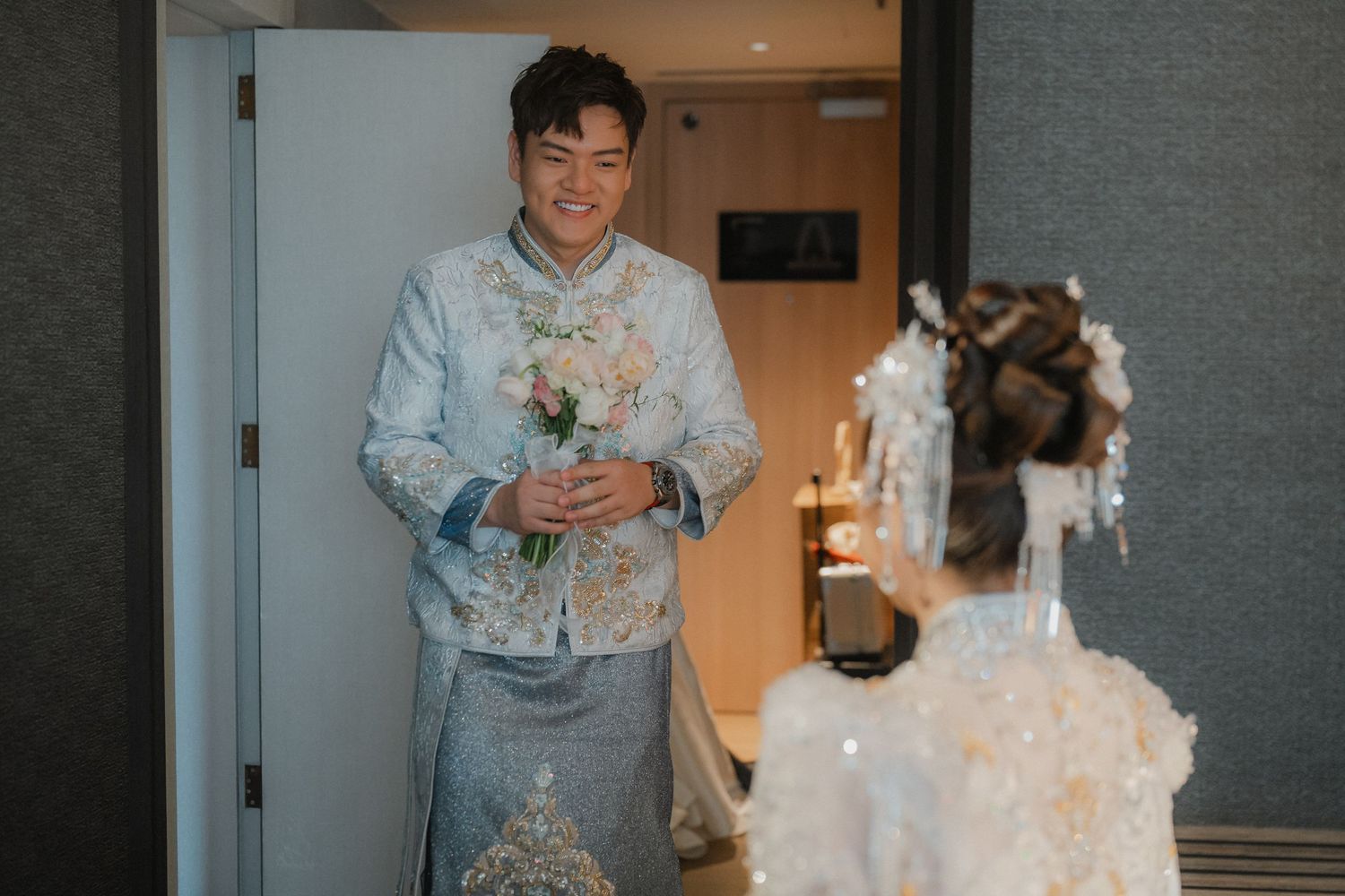 A person in a light blue floral robe holds a white coffee mug while standing against a gray wall.