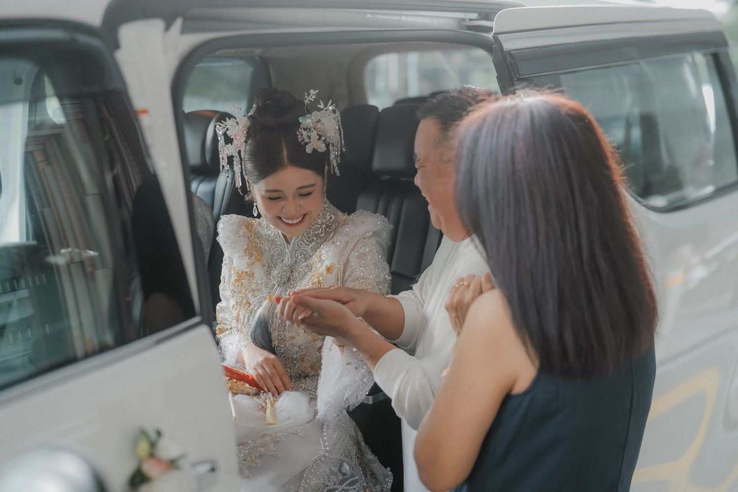 Wedding guests laugh and interact beside a white vehicle in a parking area.