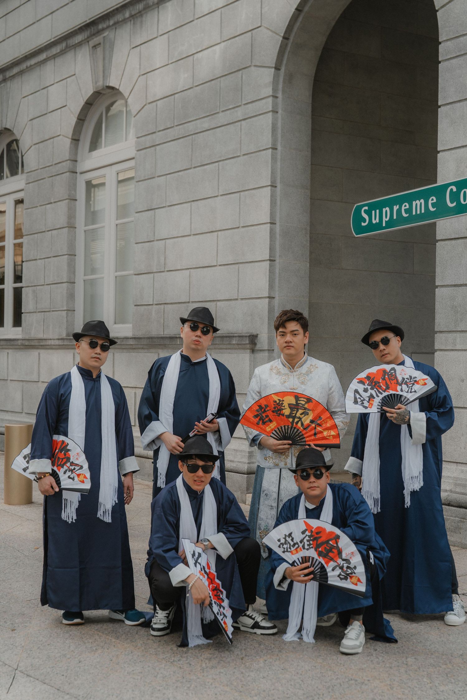 A group wearing traditional Asian robes and baseball caps poses with decorative fans under a Supreme Court street sign.