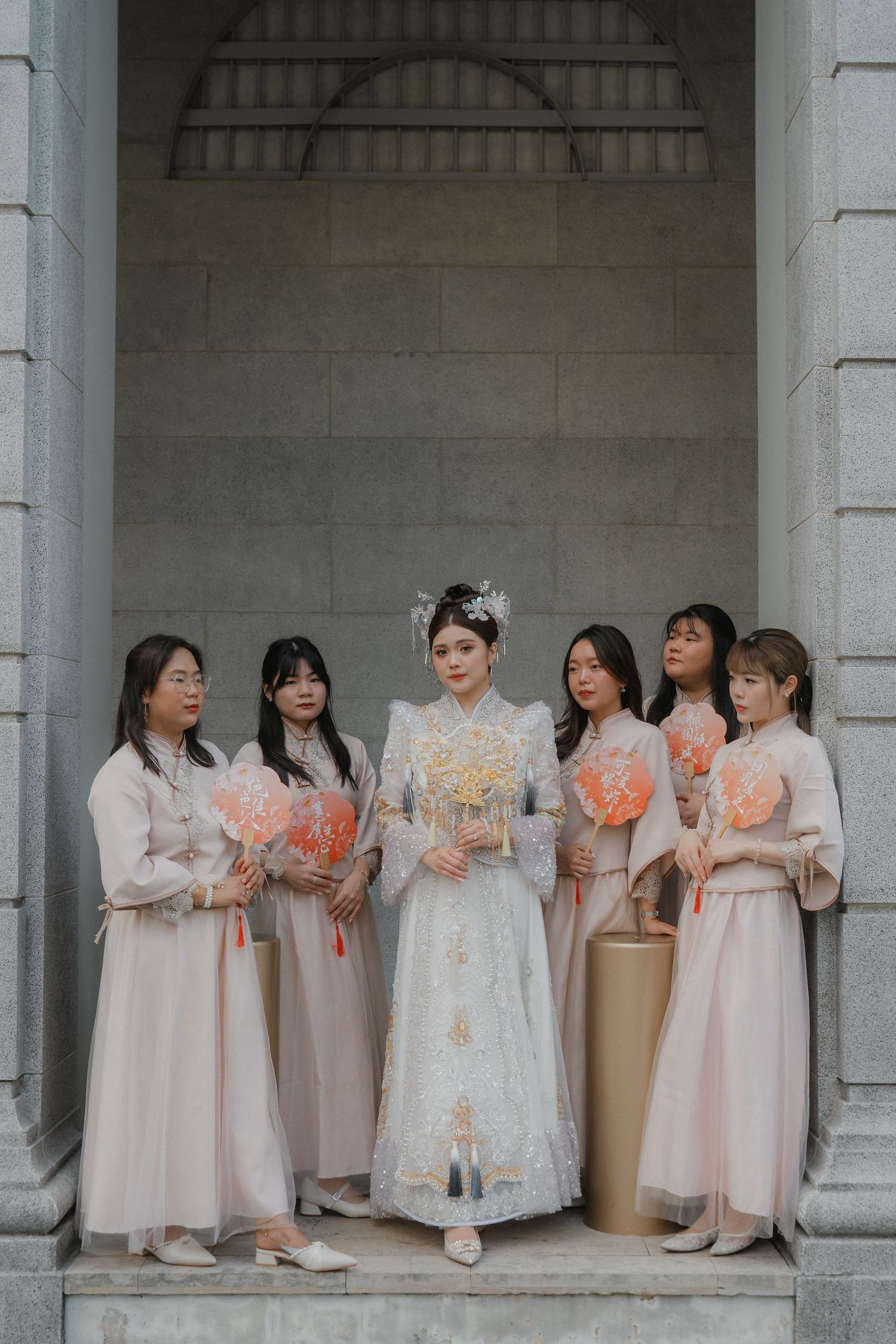 A group wearing matching light pink traditional dresses stand together in an elegant columned walkway.