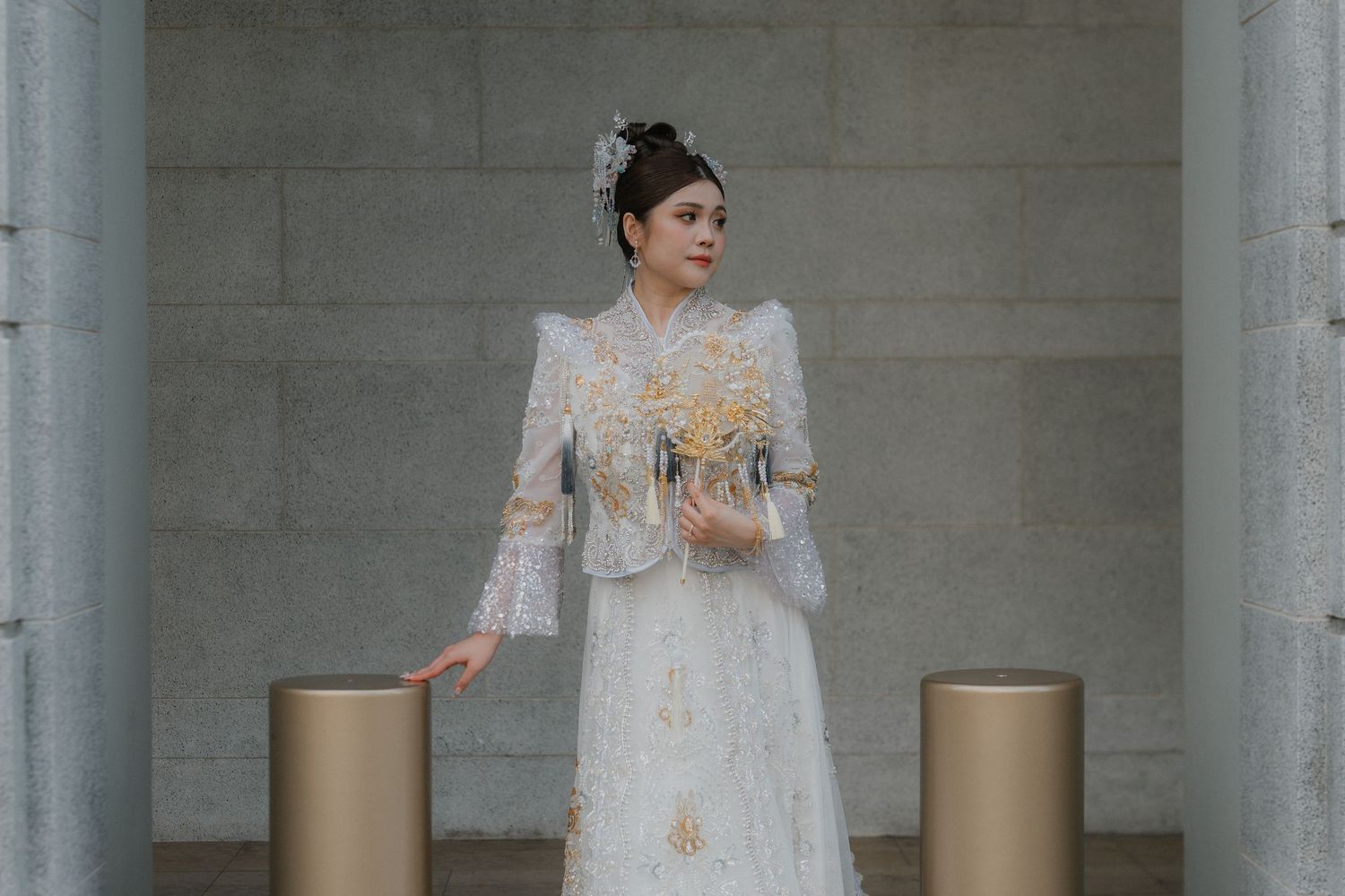 A bride in an elegant white lace gown with beaded details and bell sleeves stands between two gold pillars.