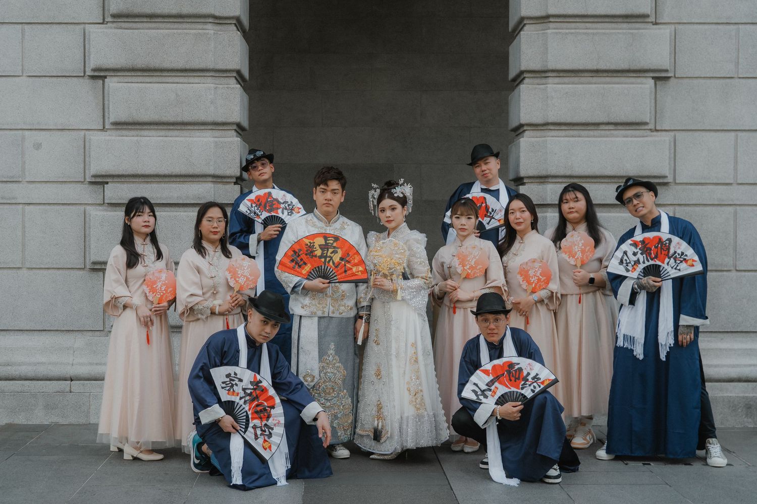 Wedding party in pale pink dresses and navy suits pose with floral bouquets against stone building exterior.