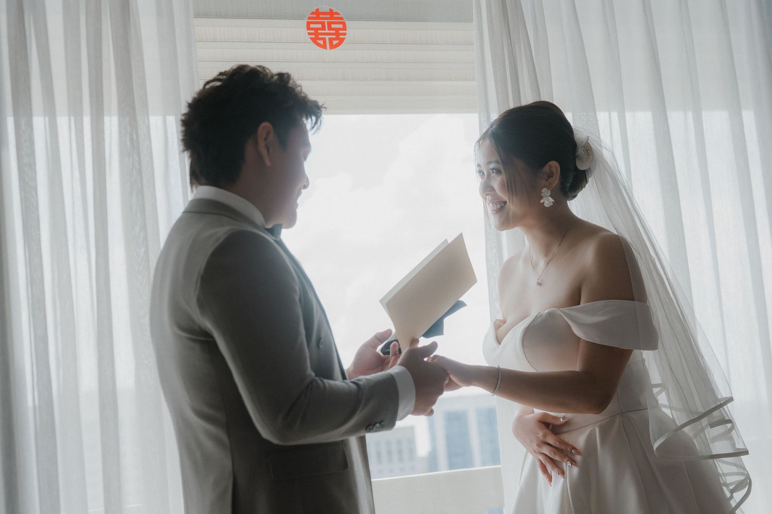 A black and white photo shows a wedding couple and guests walking through a light-filled corridor with white curtains.