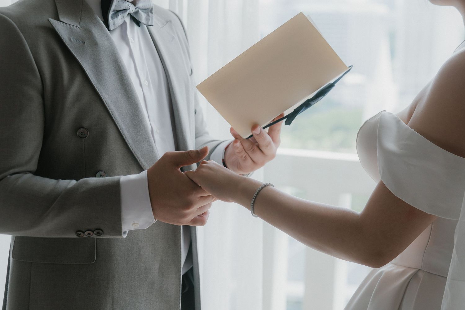 Two people wearing formal attire exchange a document between them near a bright window.