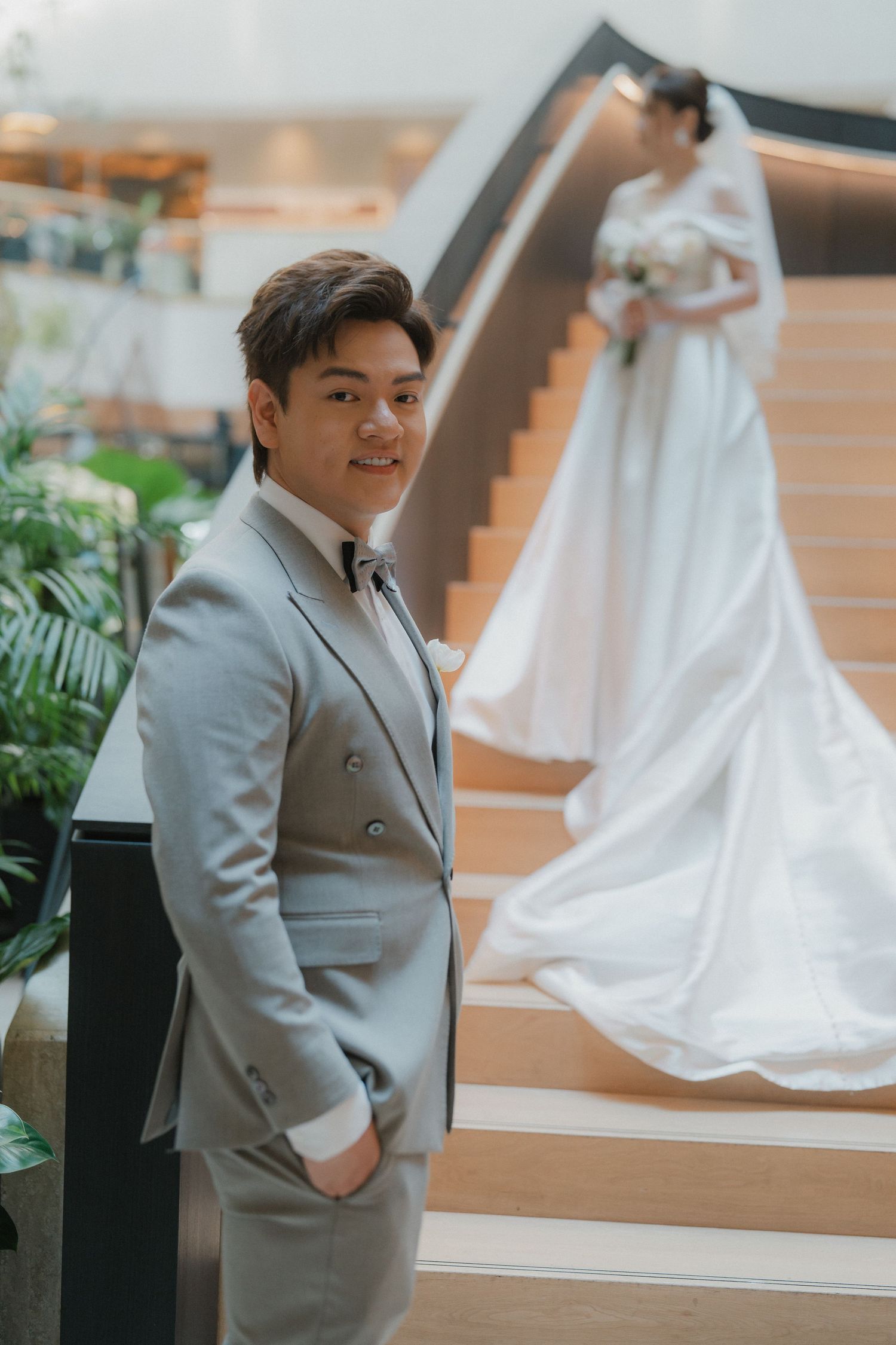 A wedding photo showing a groom in a light grey suit standing by stairs while a bride in white poses in the background.