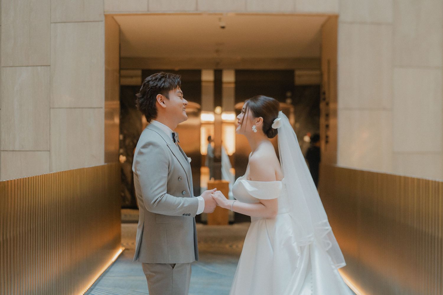 A romantic wedding scene of a couple in a warmly lit indoor corridor, with bride in white dress and groom in gray suit.
