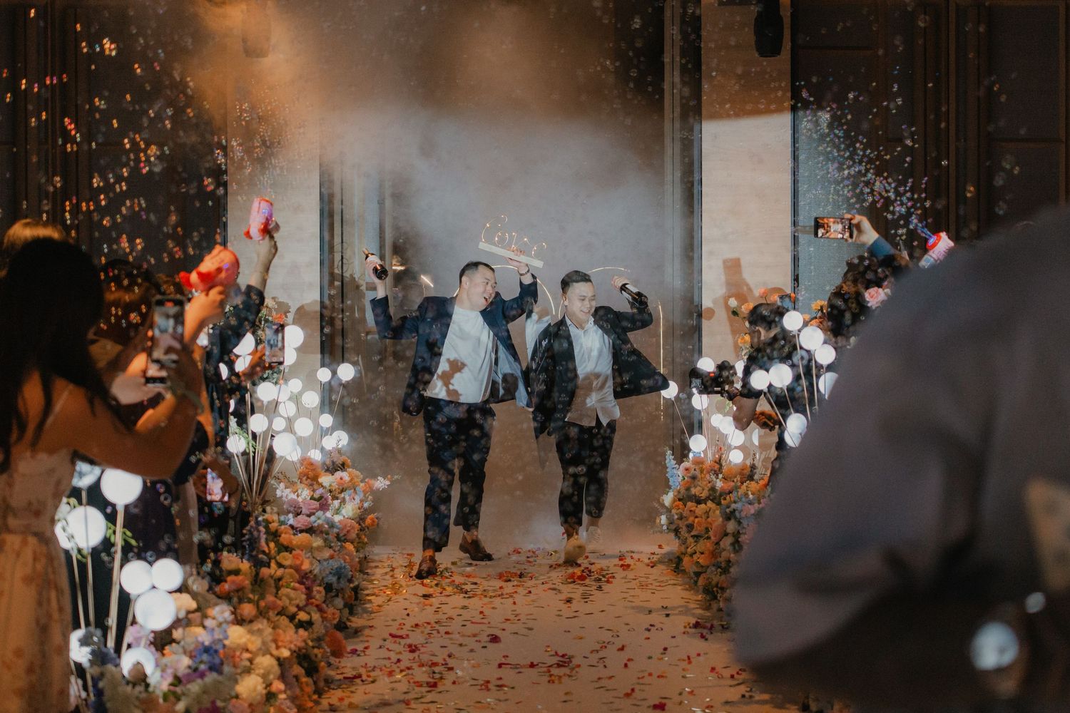 Guests line a pathway decorated with illuminated flowers as smoke fills the atmospheric space.