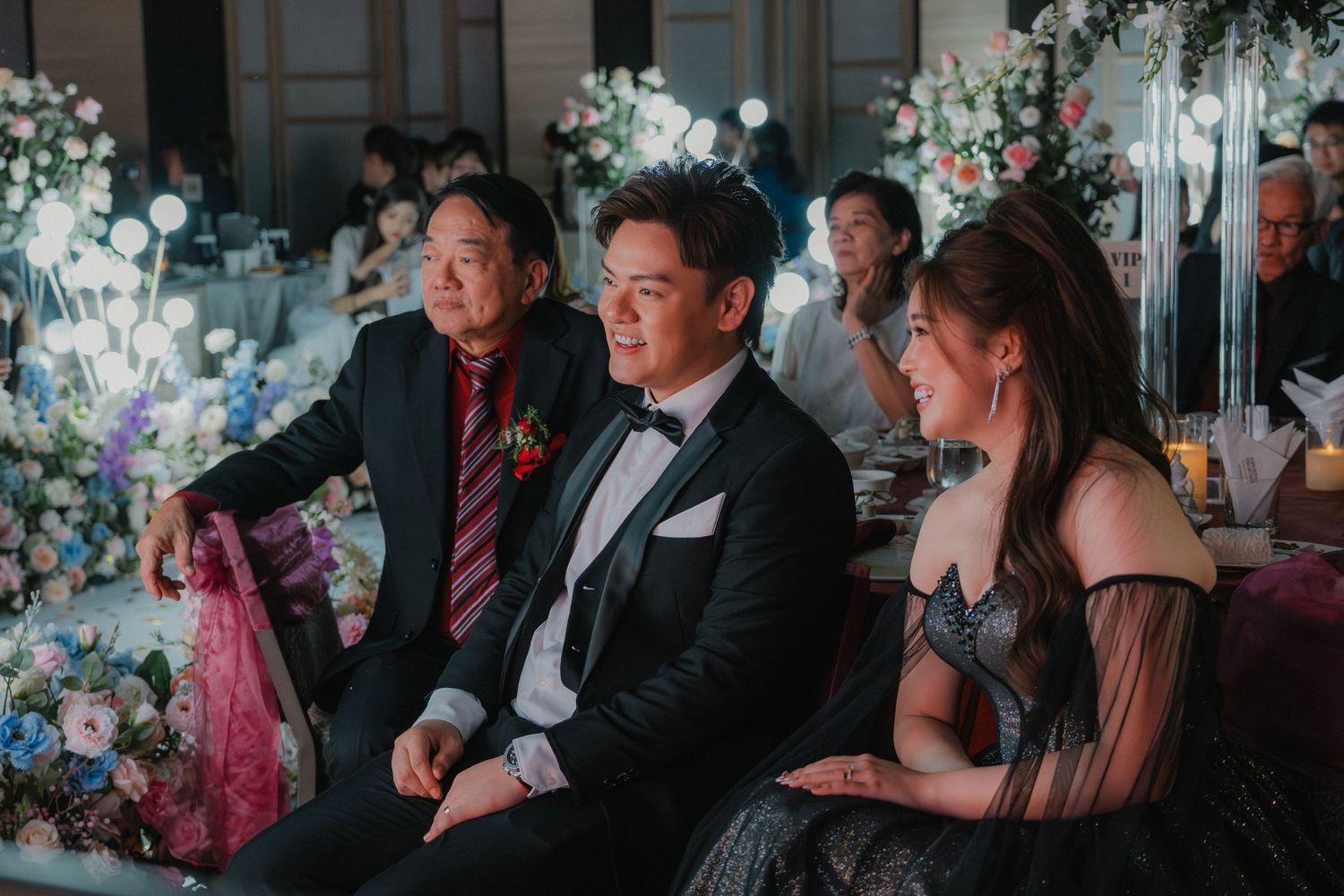 Wedding reception guests seated among elegant floral decorations in an indoor venue.