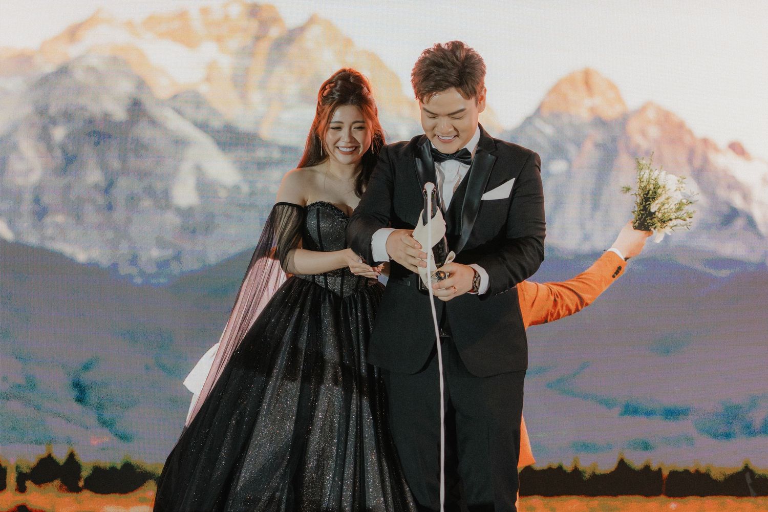 A couple in formal black attire poses against a scenic mountain backdrop at sunset for a wedding photoshoot.