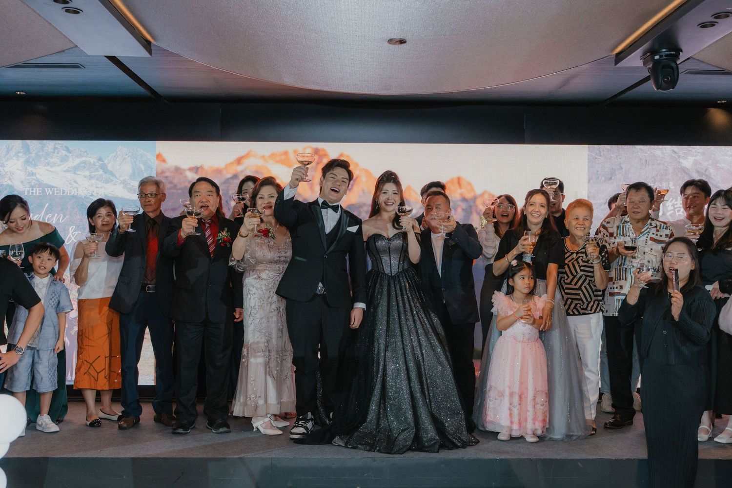 Large group wedding photo with couple and guests in formal attire against dark background.