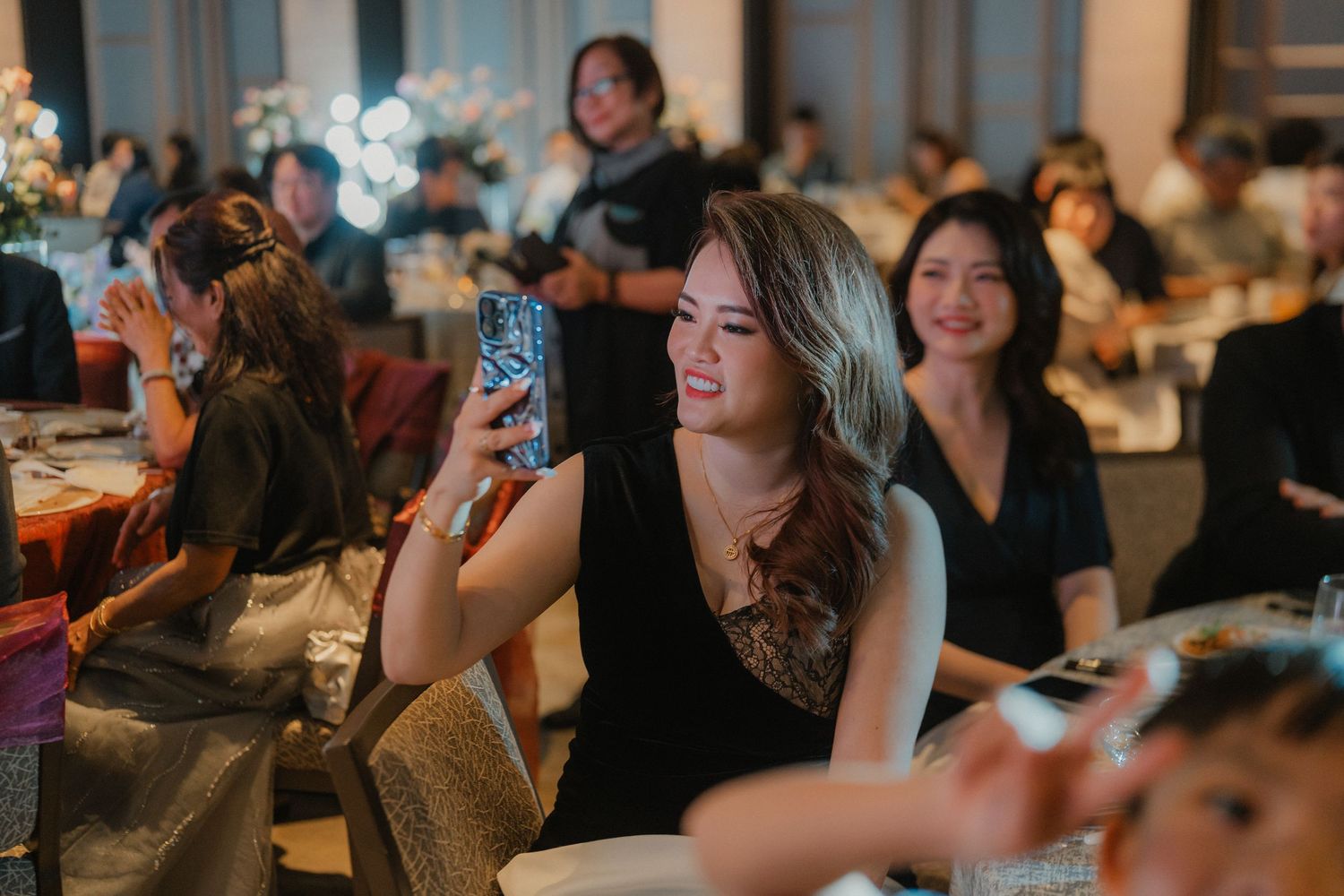 Wedding guests in a dimly lit reception venue smile and take photos during the celebration.