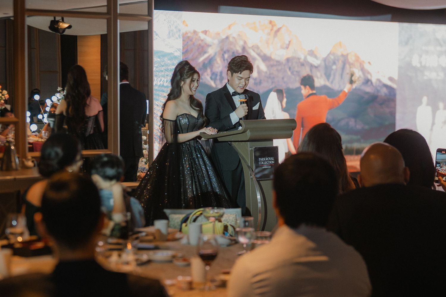 A couple gives a speech at an elegant wedding reception with mountain scenery projected on a wall behind them.