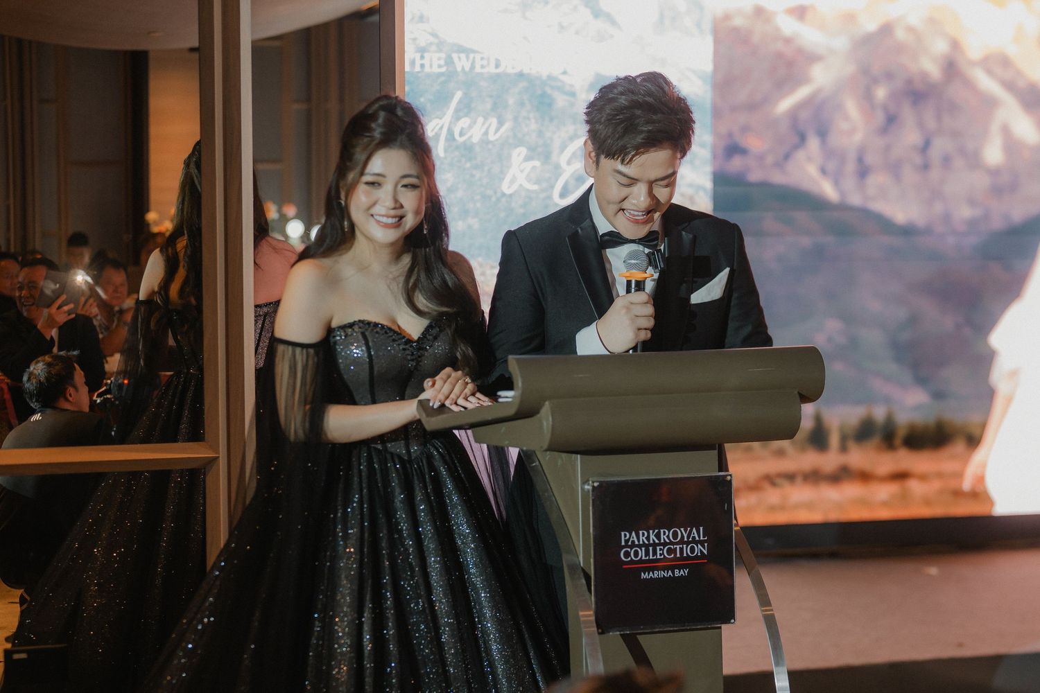 A couple in black formal attire presents at a podium with mountain scenery displayed behind them.