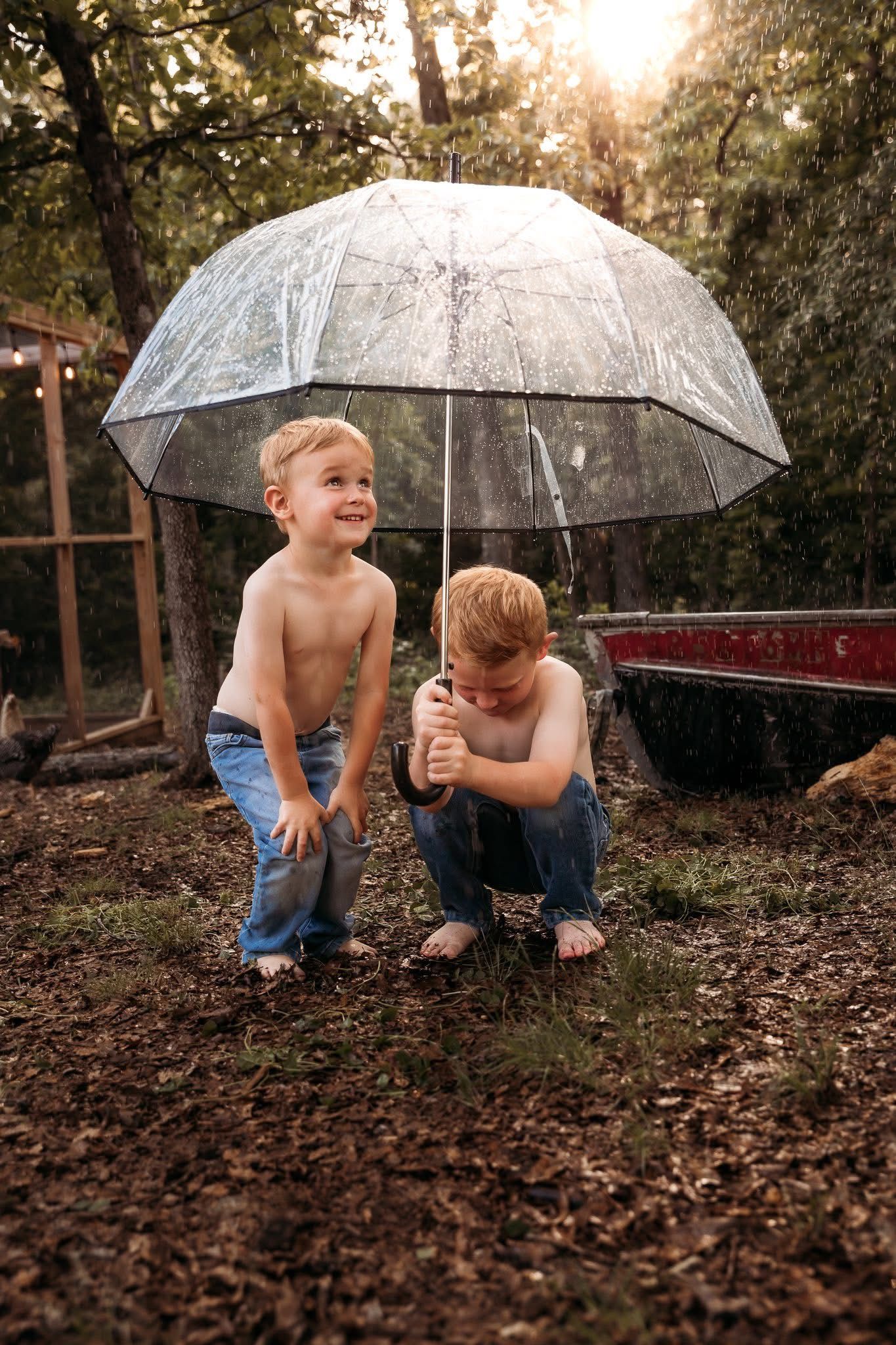 Two young boys sharing clear umbrella outdoors at sunset.