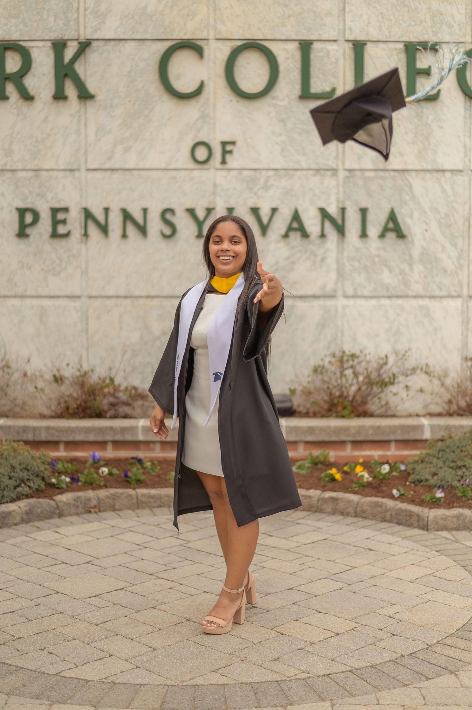 Graduate in cap and gown poses creatively against beige wall.