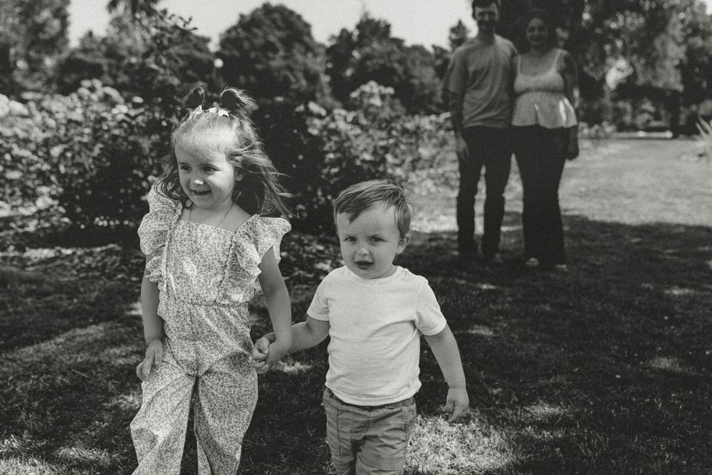Two young siblings hold hands and walk through a lush garden in black and white photo.