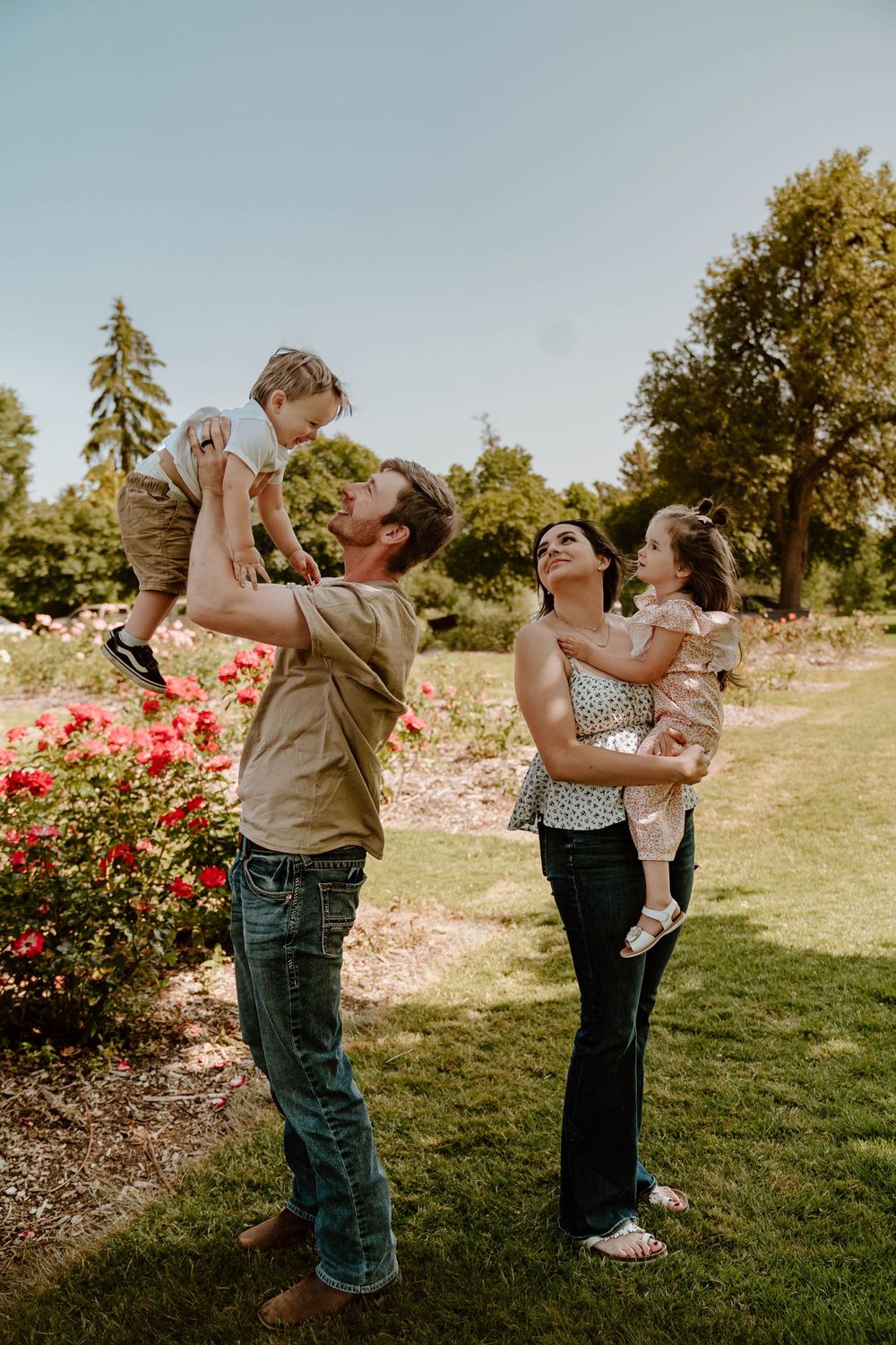 Joyful parents lift their two young children in a colorful rose garden on a sunny day.