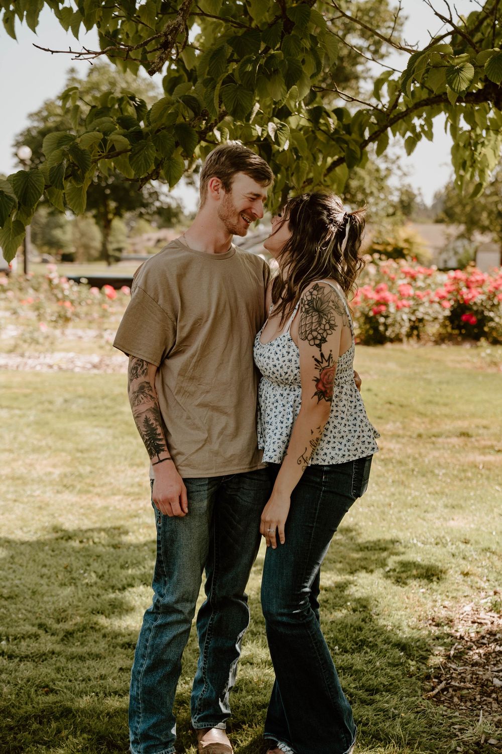 Smiling tattooed couple share a romantic moment under a tree with roses in the background.