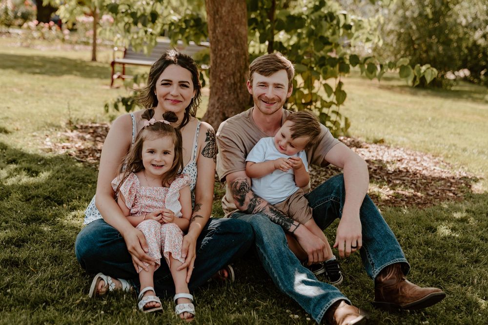 Happy family of four sits together on green grass in a sunny garden setting.