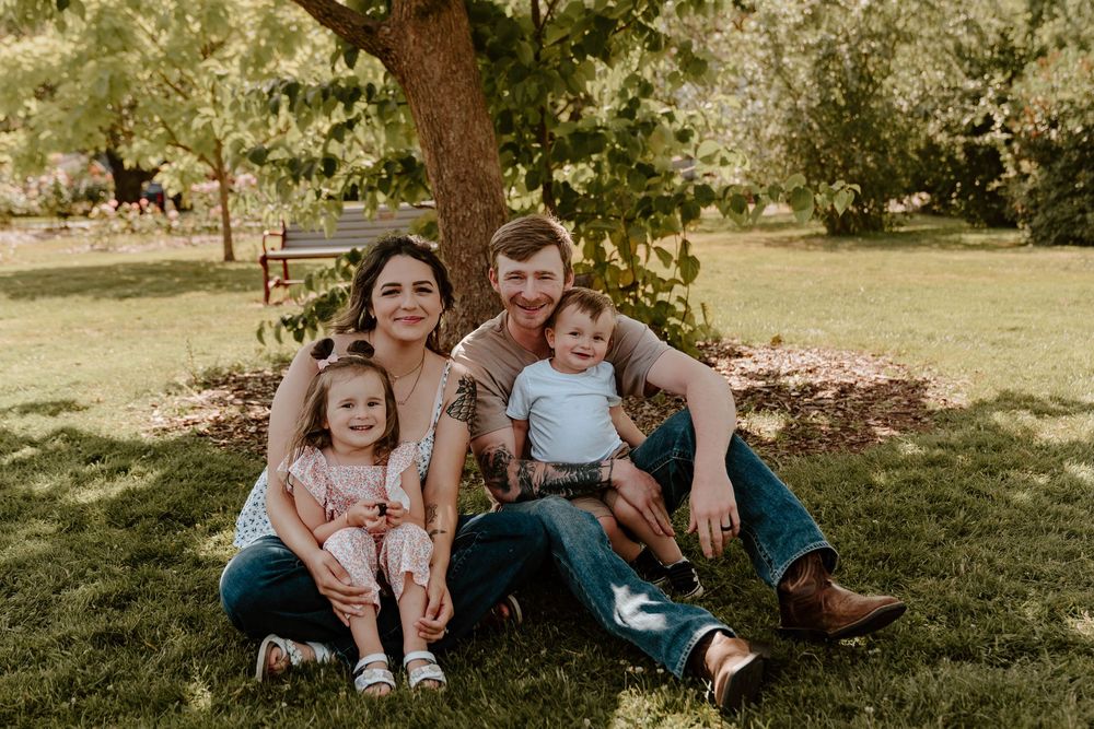 Smiling family of four sits casually on green grass beneath a shady tree outdoors.