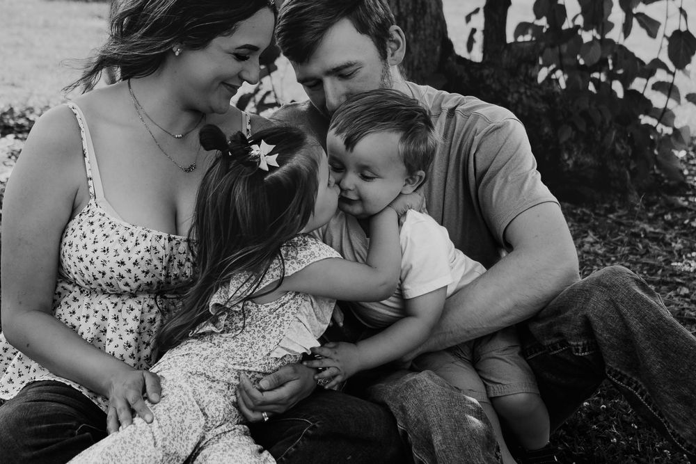 Tender black and white family portrait of parents and two young children sitting under a tree.