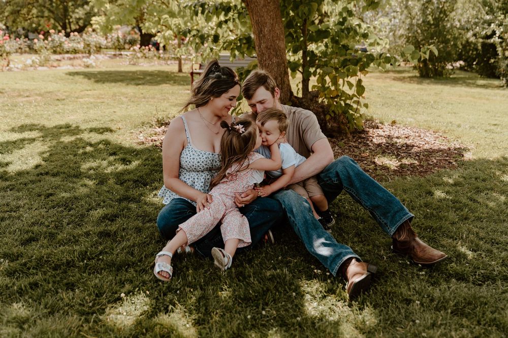 Happy family of four laughs and cuddles together on green grass beneath a shady tree.
