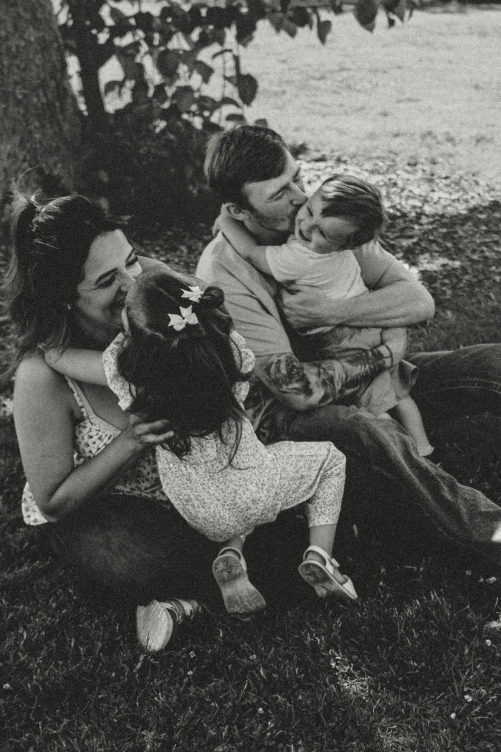 Candid black and white family photo of parents and children sharing joyful hugs under a tree.