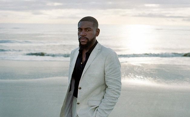 A person in a white blazer and black shirt walks along a serene beach at sunset creating multiple scenic poses.