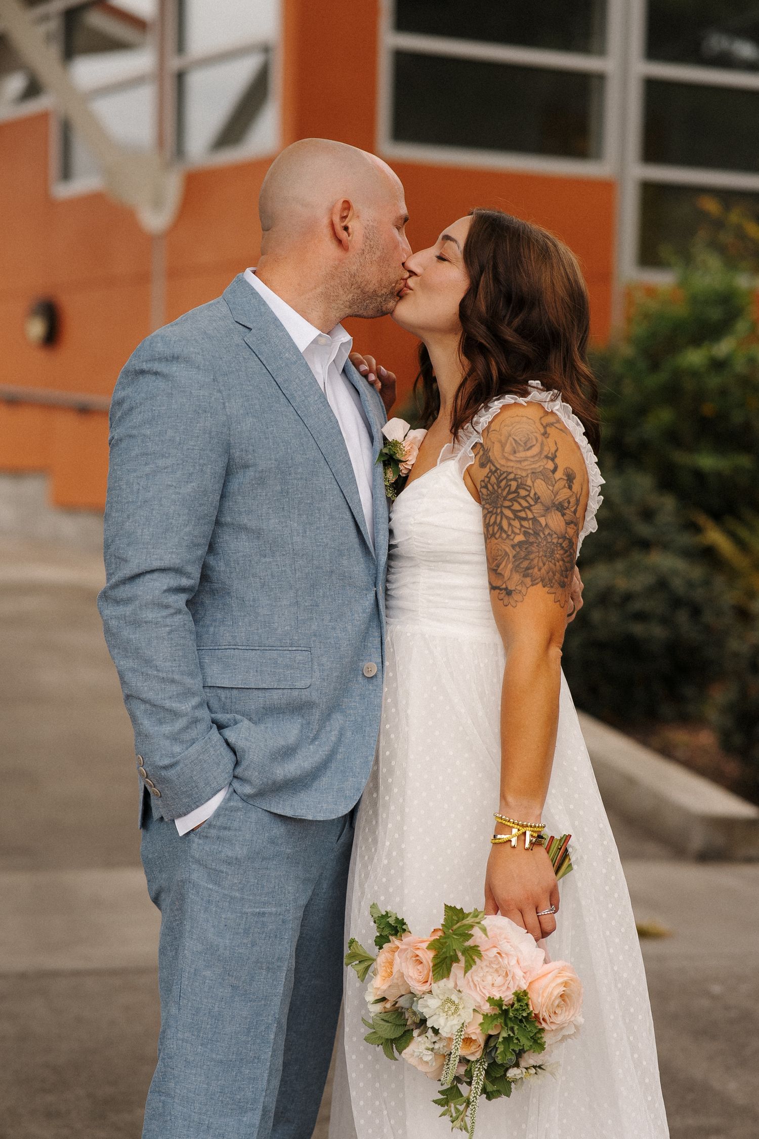 A couple shares an intimate kiss outdoors, with the bride holding a peachy floral bouquet against her white polka dot dress.