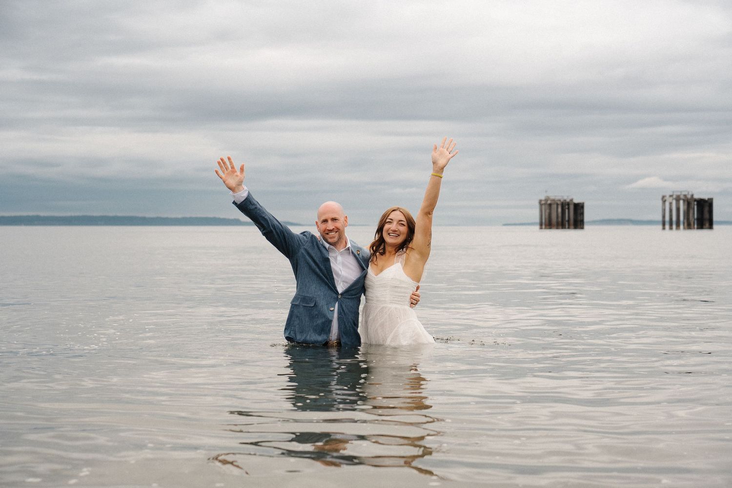 A couple in formal wedding attire raises their arms joyfully while standing in calm lake waters with mountains in background.