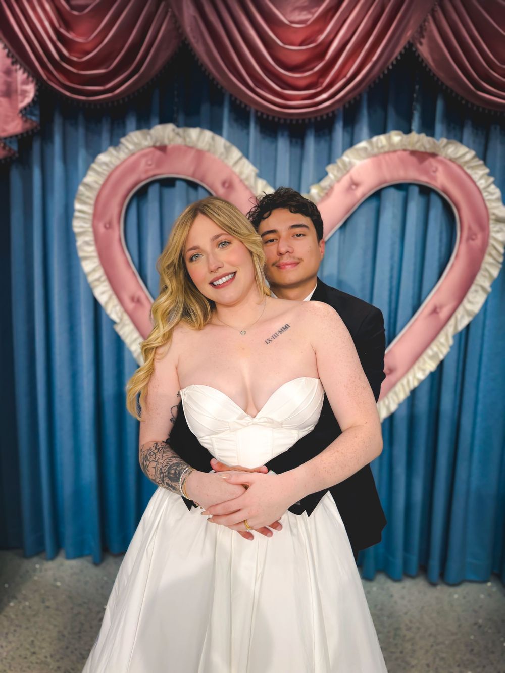 Two people in white wedding attire pose together in front of a blue curtain with pink heart decorations.