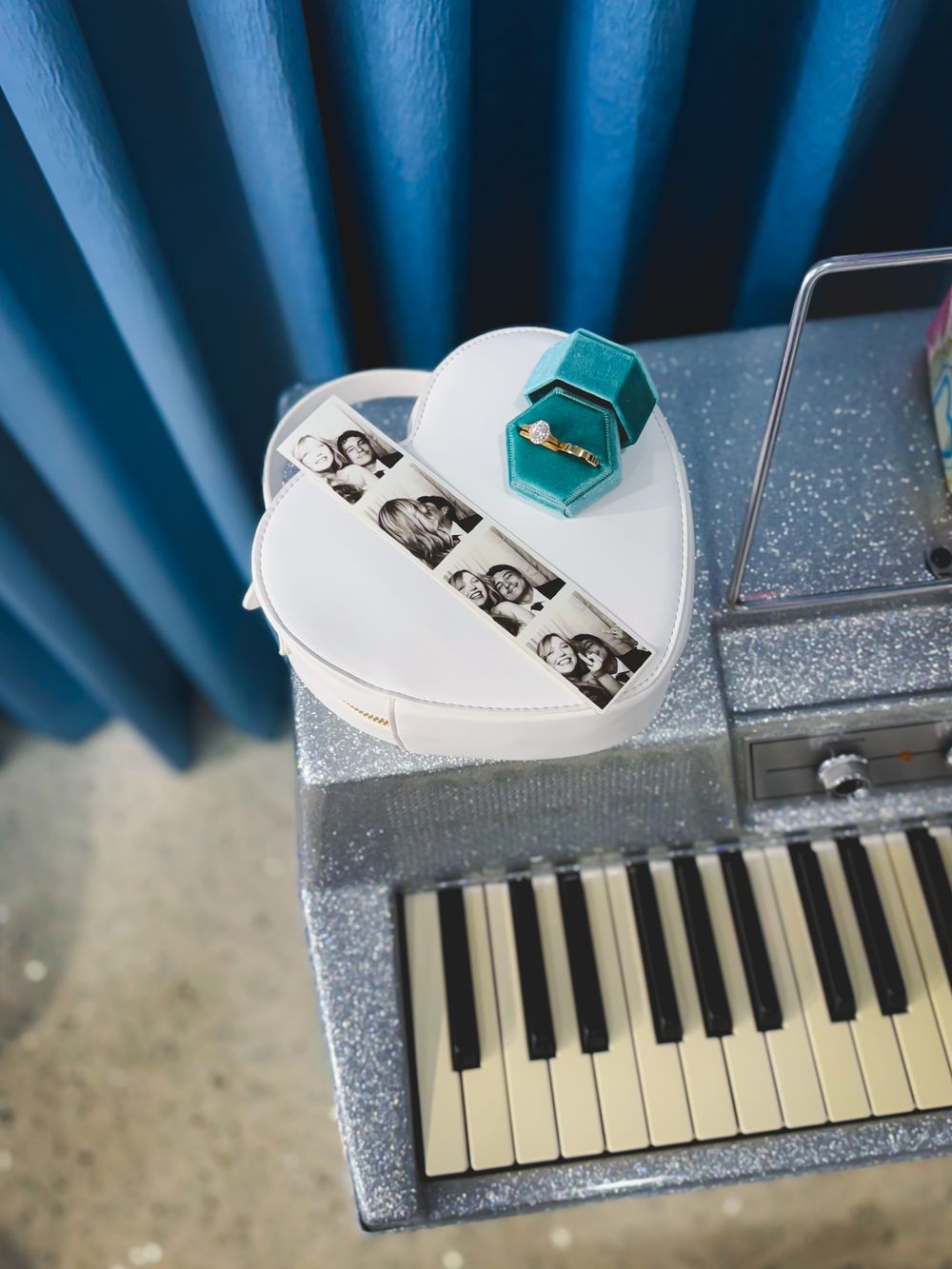 A white heart-shaped box with black photo frames sits atop a silver sparkly keyboard or synthesizer with blue curtains behind.