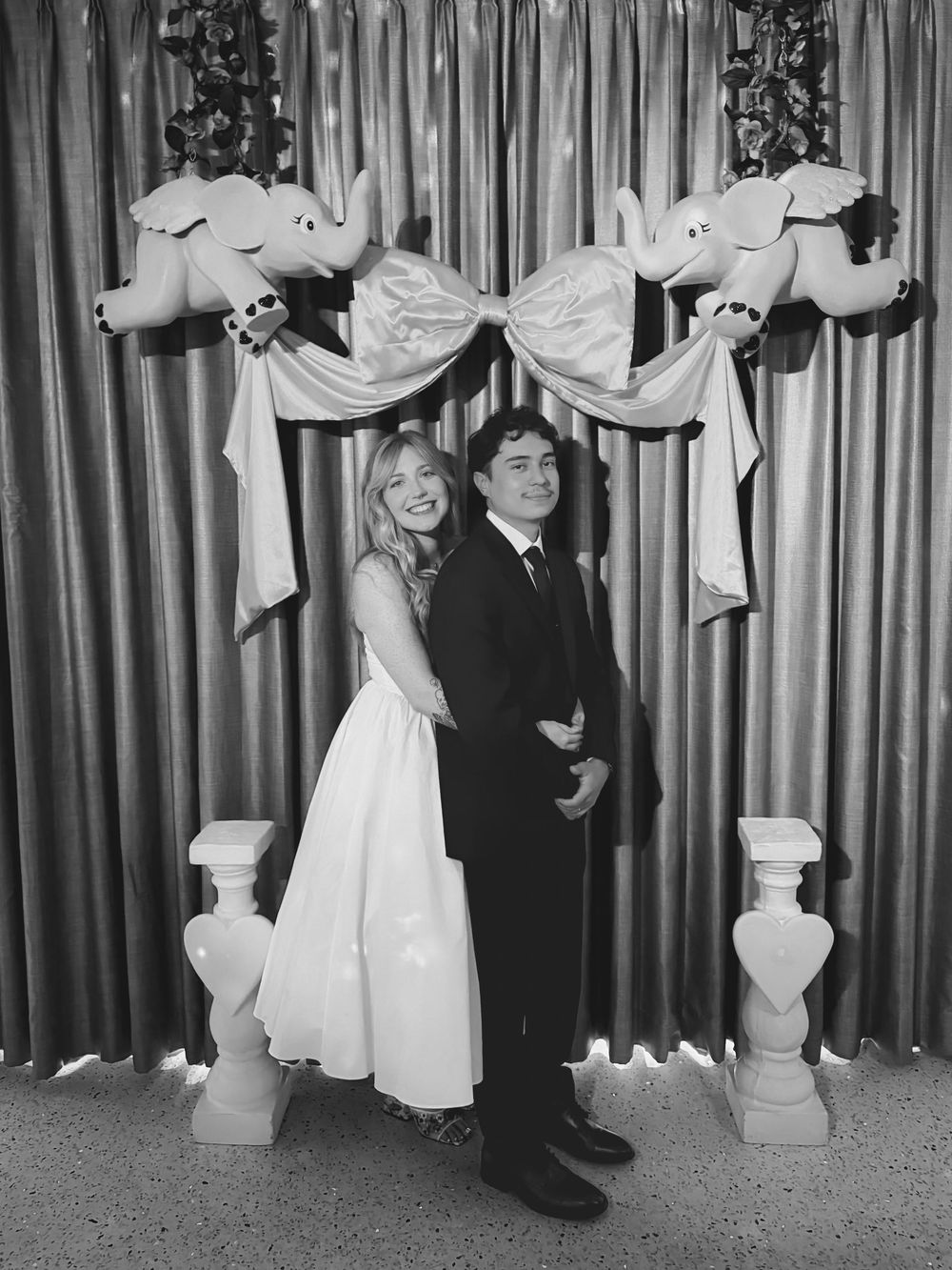Black and white photograph of a high school dance couple posing under decorative white garlands and columns at a formal event.