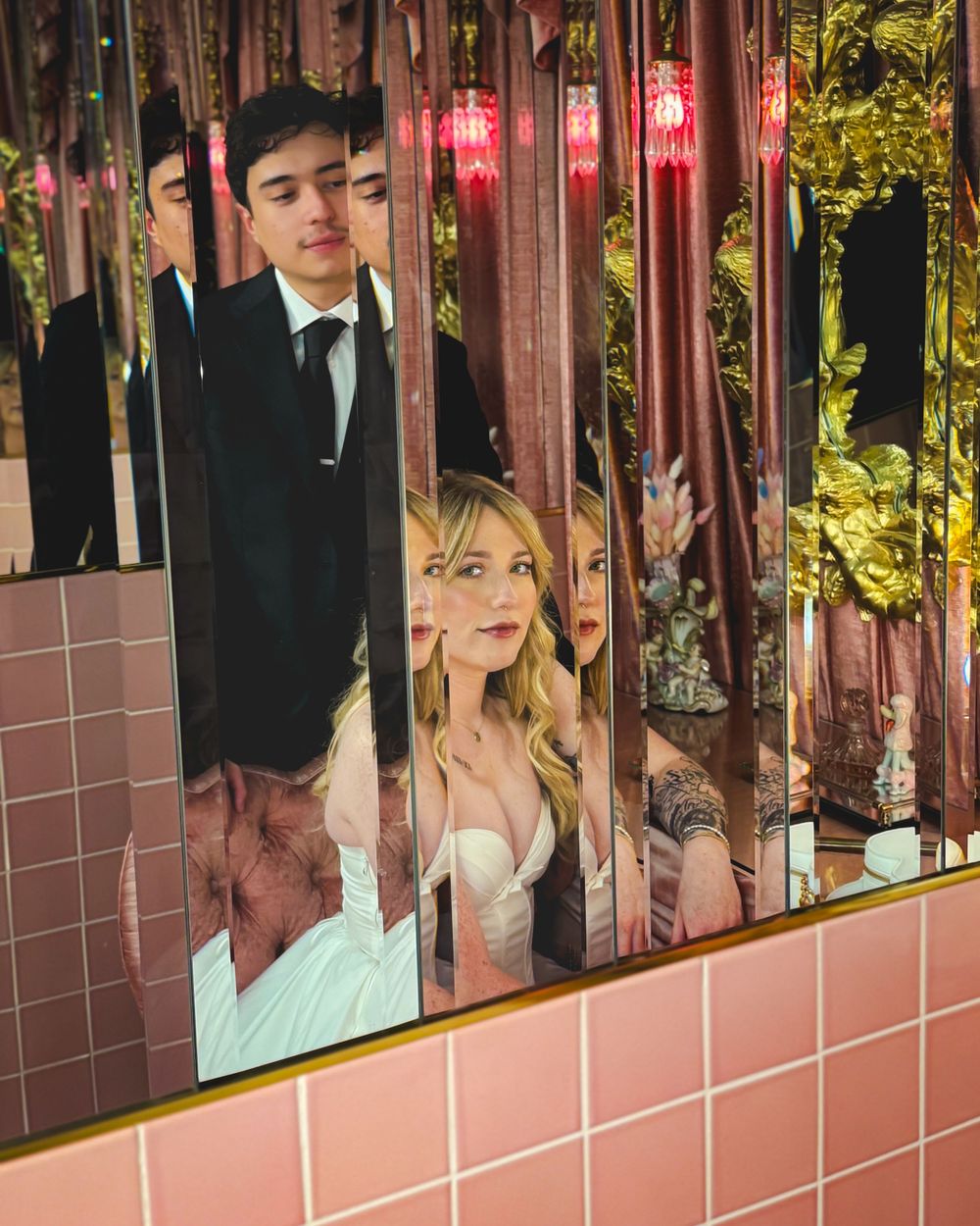 Two people pose together at a pink-tiled venue with decorative metalwork and green foliage visible in the background.