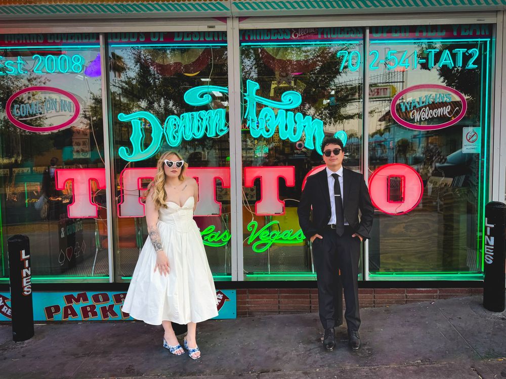 Bride and groom pose outside vintage-style tattoo shop with neon signage and retro storefront decor.
