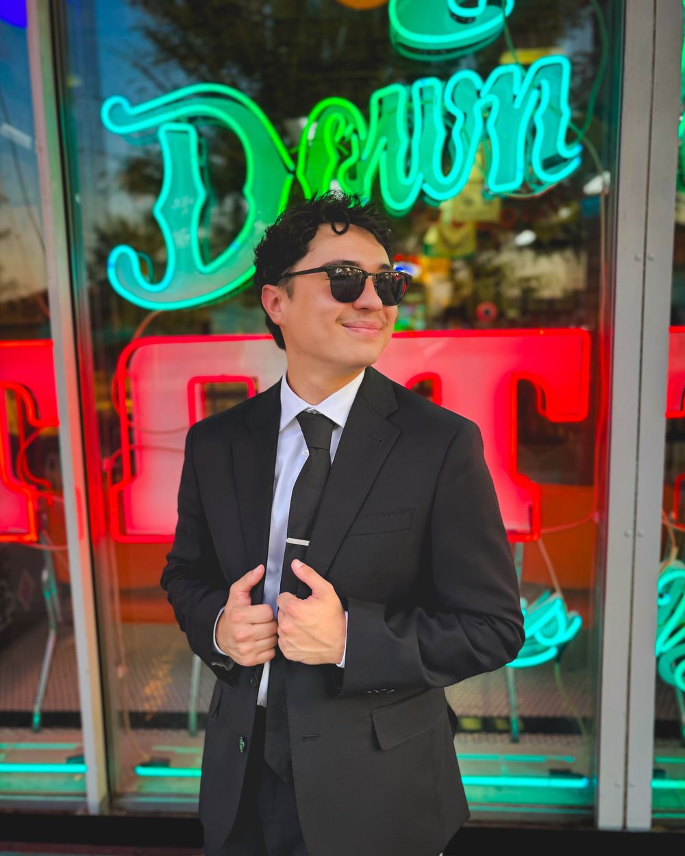 A well-dressed person in a black suit and tie poses confidently in front of a neon Dream sign.