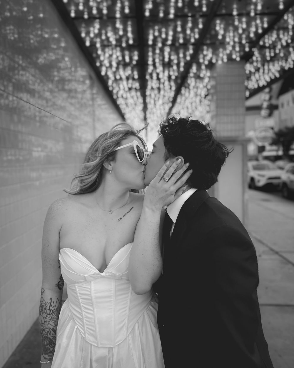 A romantic black and white wedding photo shows a couple sharing a kiss under twinkling lights on an urban street.