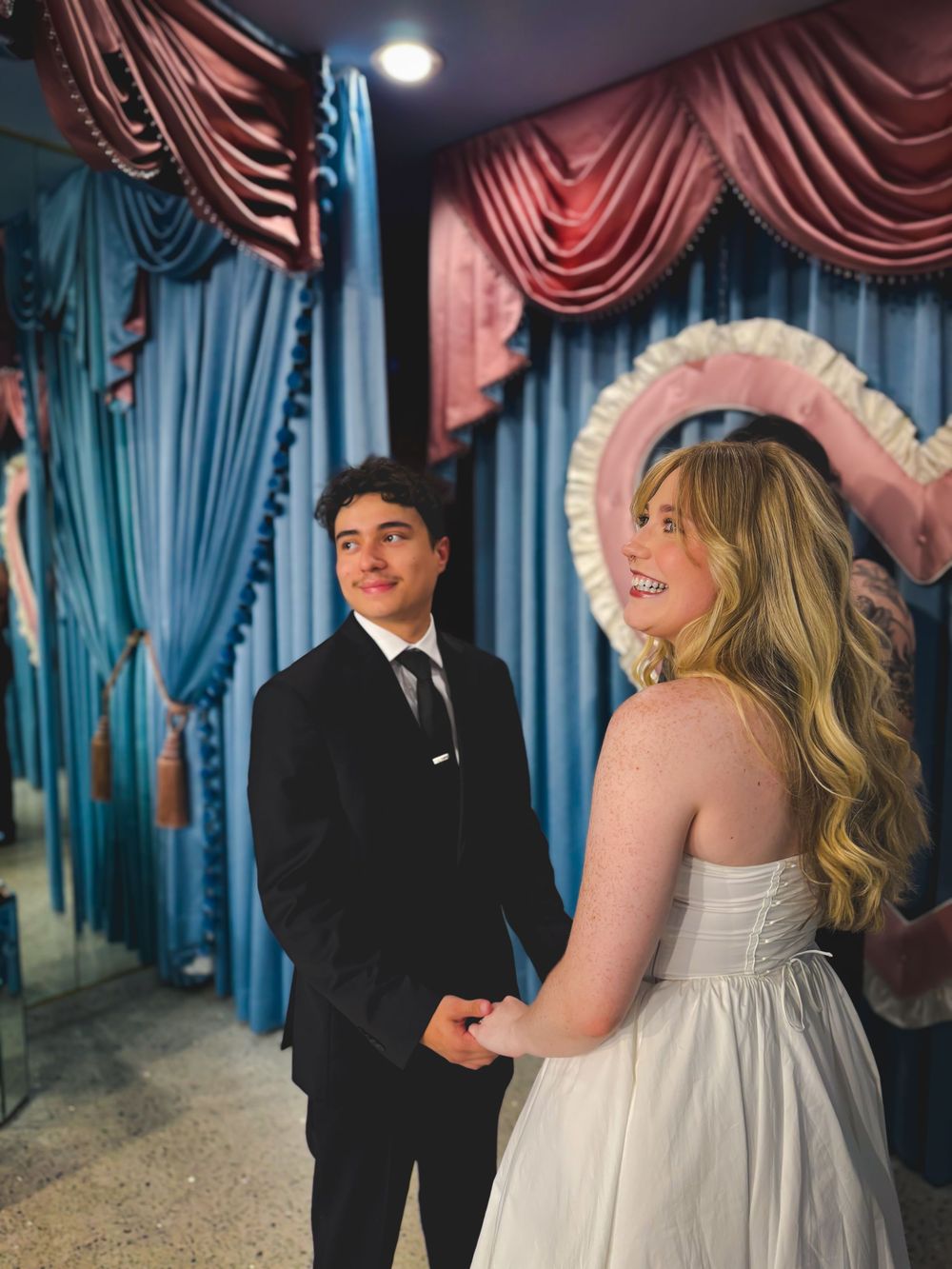 A couple in formal wedding attire share a romantic moment against a backdrop of blue and coral draped curtains.