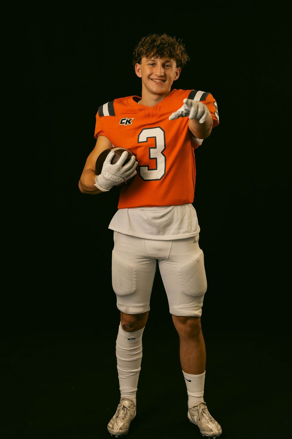 High school football player poses in orange jersey and white shorts with a football against dark background.
