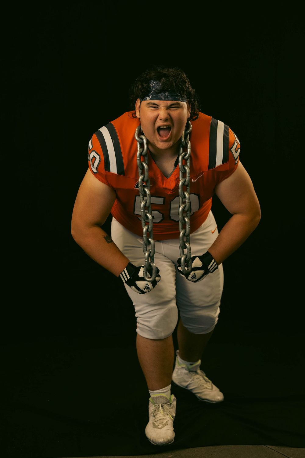 Football player in orange jersey and white pants making an intense aggressive expression against dark background.