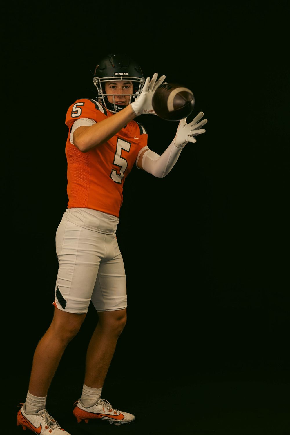 Football player in orange jersey number 5 reaching out to catch a football against black backdrop.
